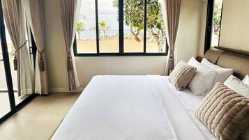 Een groot bed met witte lakens en beige kussens, een raam met gordijnen en uitzicht op een strand.