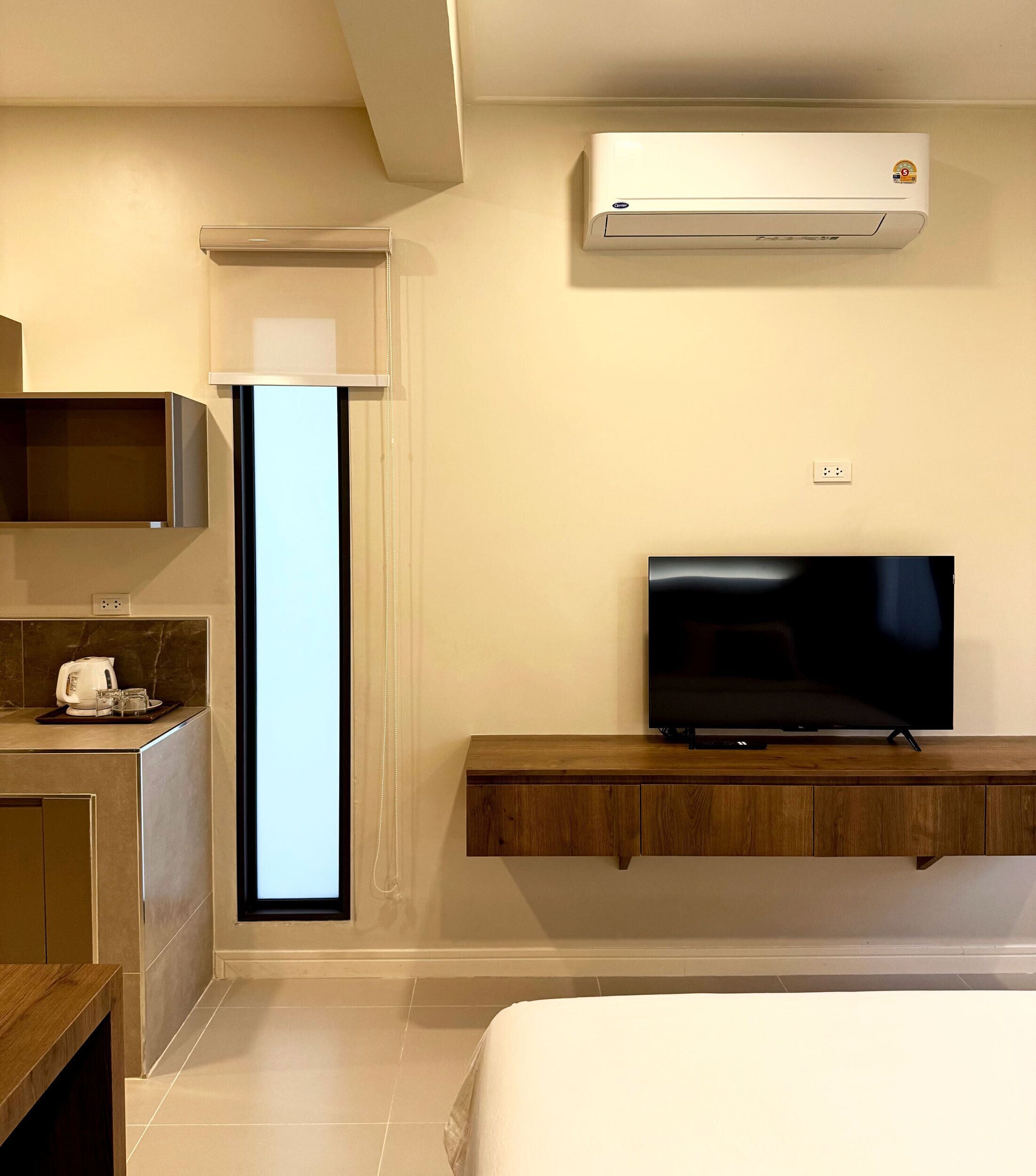 A modern hotel room with a flat-screen TV on a wooden shelf, a built-in air conditioning unit, a kitchen area with a coffee maker, and a large window.
