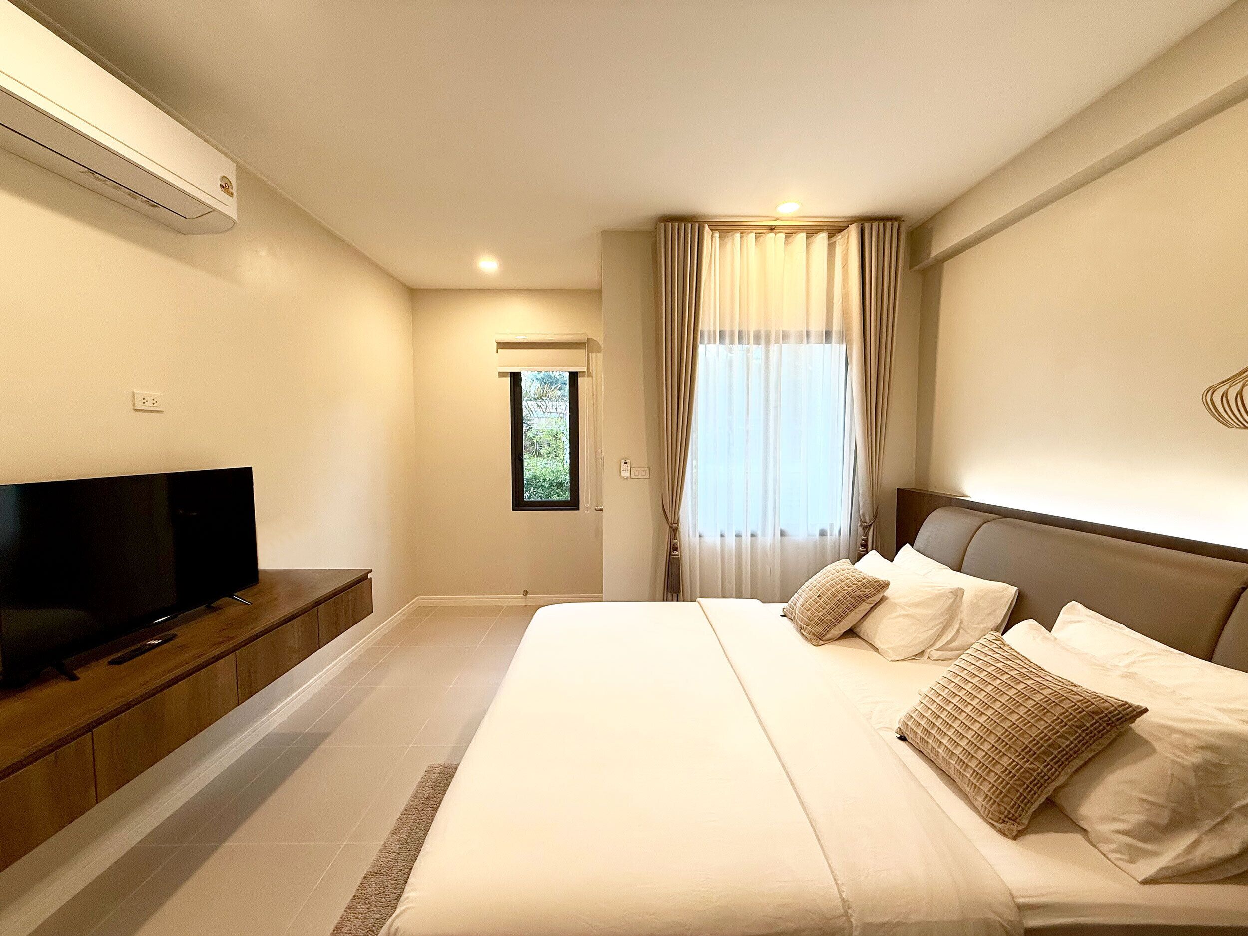 A modern hotel room with a large bed, a flat-screen TV on a wooden console, a window with sheer curtains, and a ceiling-mounted air conditioner.