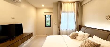 Quarto de hotel moderno com uma cama grande, uma televisão de tela plana sobre um console de madeira, uma janela com cortinas finas e um ar-condicionado instalado no teto.