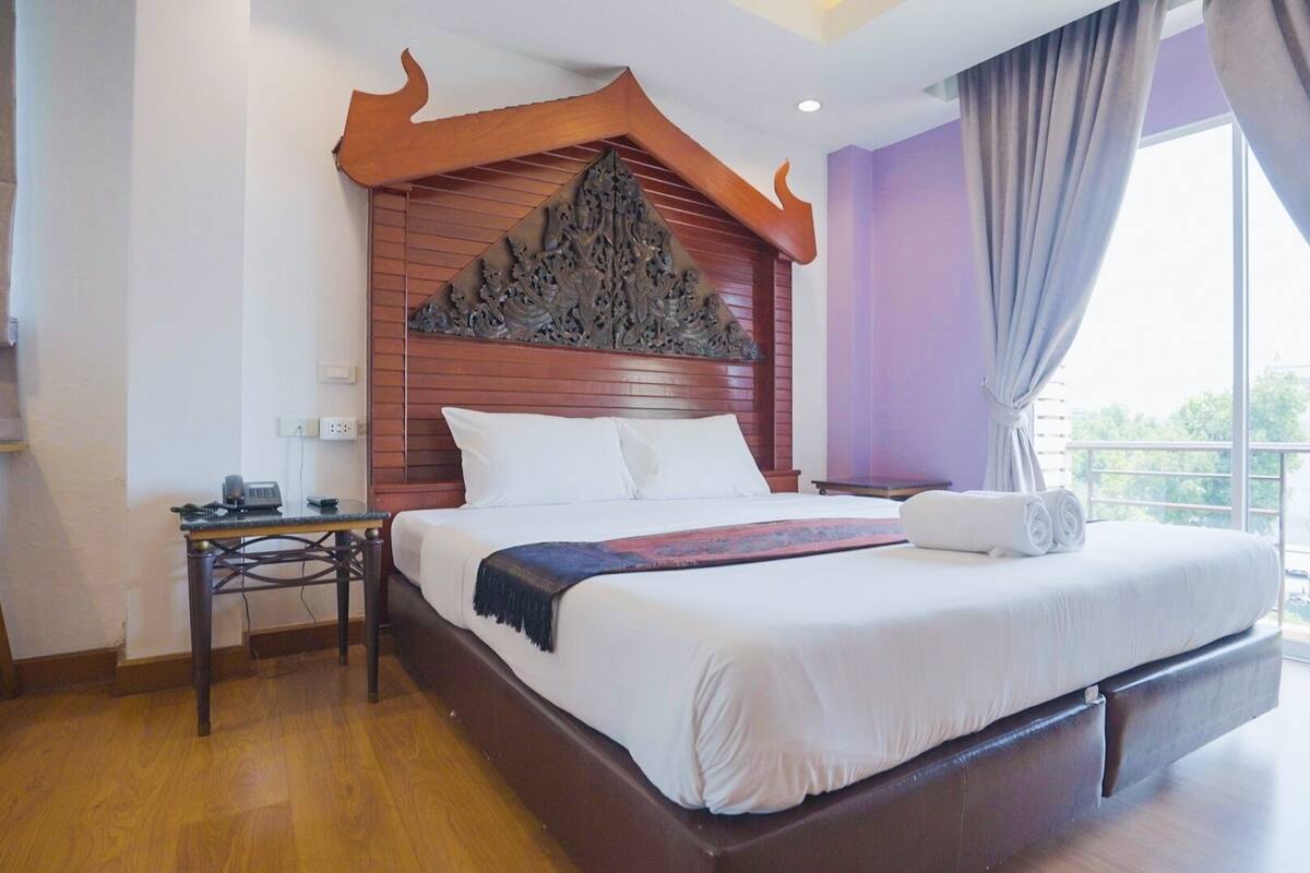 A hotel room with a large bed, a wooden headboard, a bedside table, and a view of the outside.