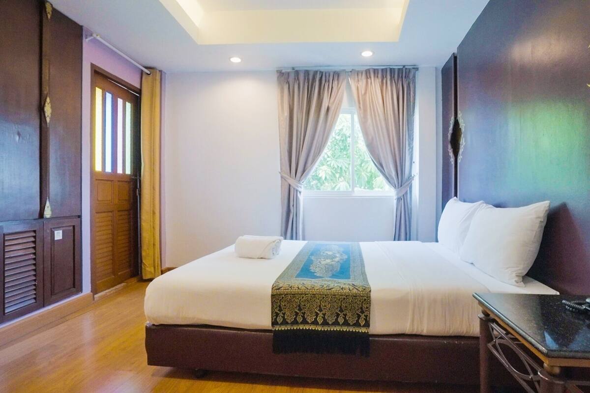 A hotel room with a large bed, a bedside table with a phone, a window with curtains, and a view of greenery outside.