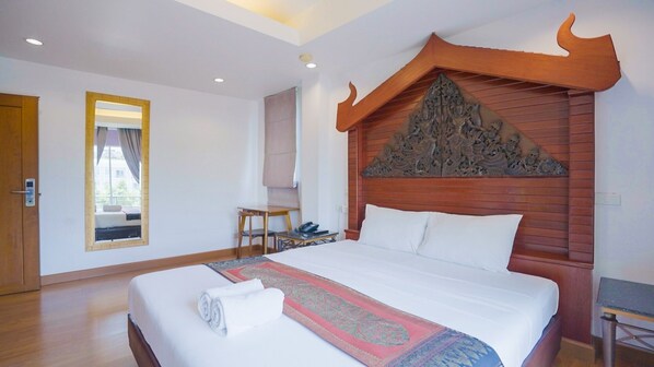 A hotel room with a large bed, a wooden headboard with intricate carvings, a small desk with a chair, and a window with curtains. - Korbua House (Bangkok)