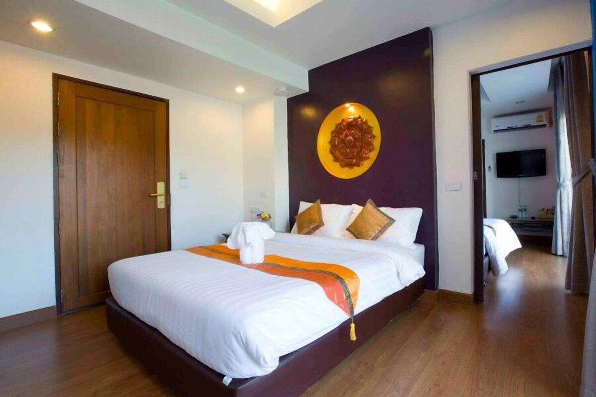A hotel room with a large bed, a wooden door, a wall-mounted TV, and a hallway leading to another room.