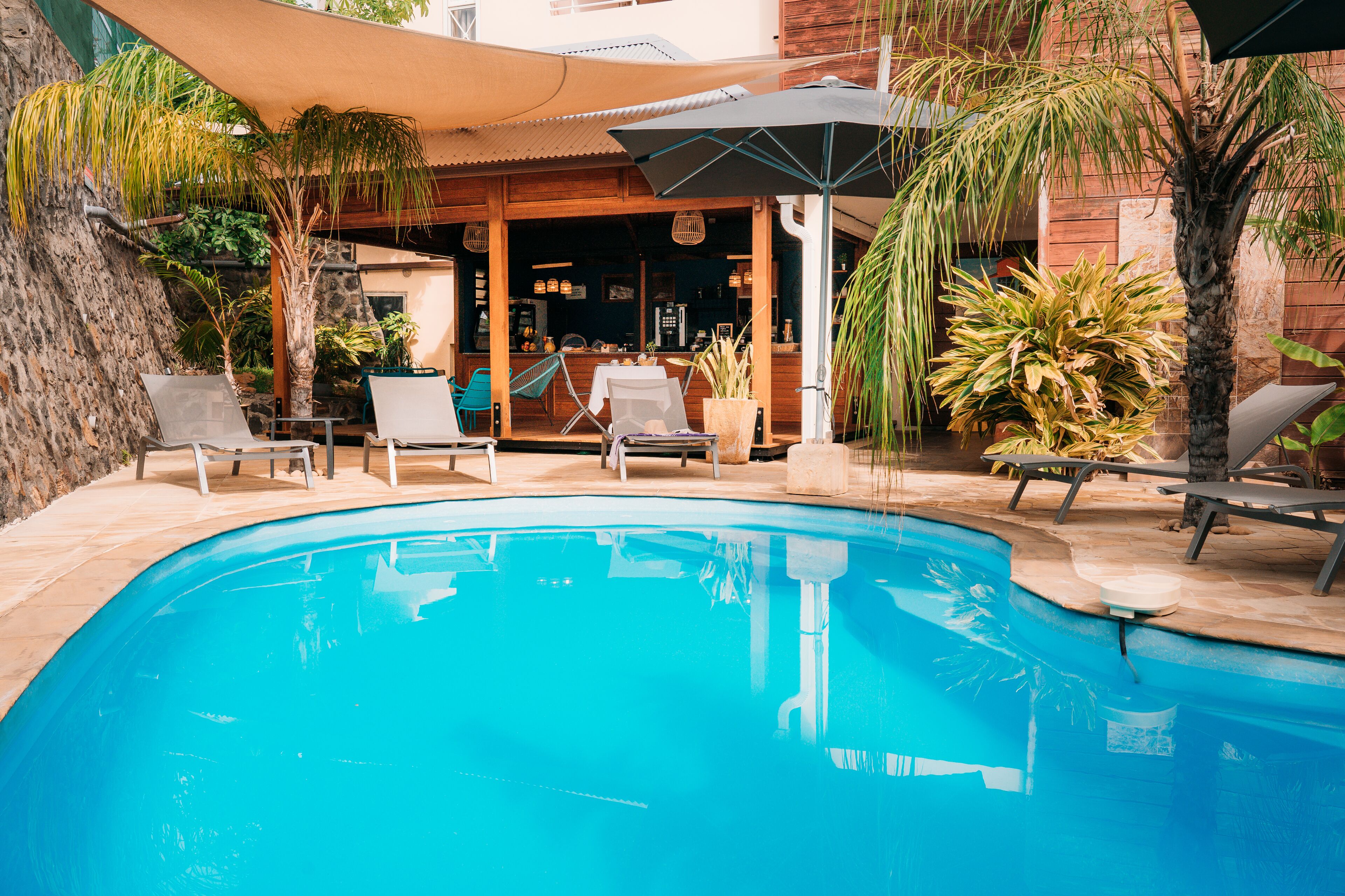 A swimming pool with lounge chairs and umbrellas, surrounded by plants and a building with a bar area.