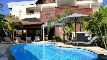 Una piscina con sillones de descanso y sombrillas, rodeada por un edificio con balcón y plantas en macetas.