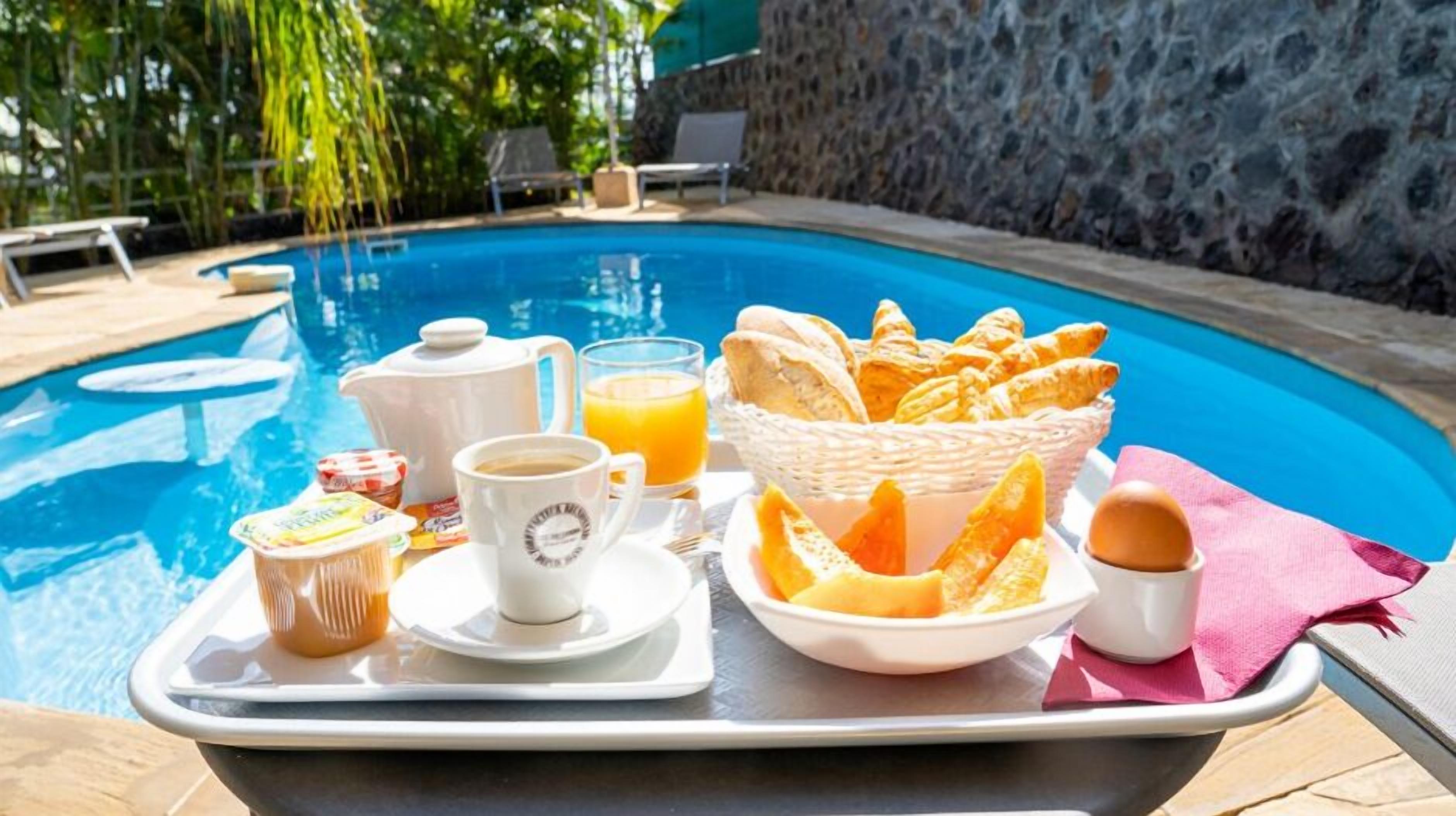 Bandeja de desayuno con cruasanes, zumo de naranja, café y yogur junto a una piscina.