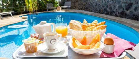 Bandeja de desayuno con cruasanes, zumo de naranja, café y yogur junto a una piscina.