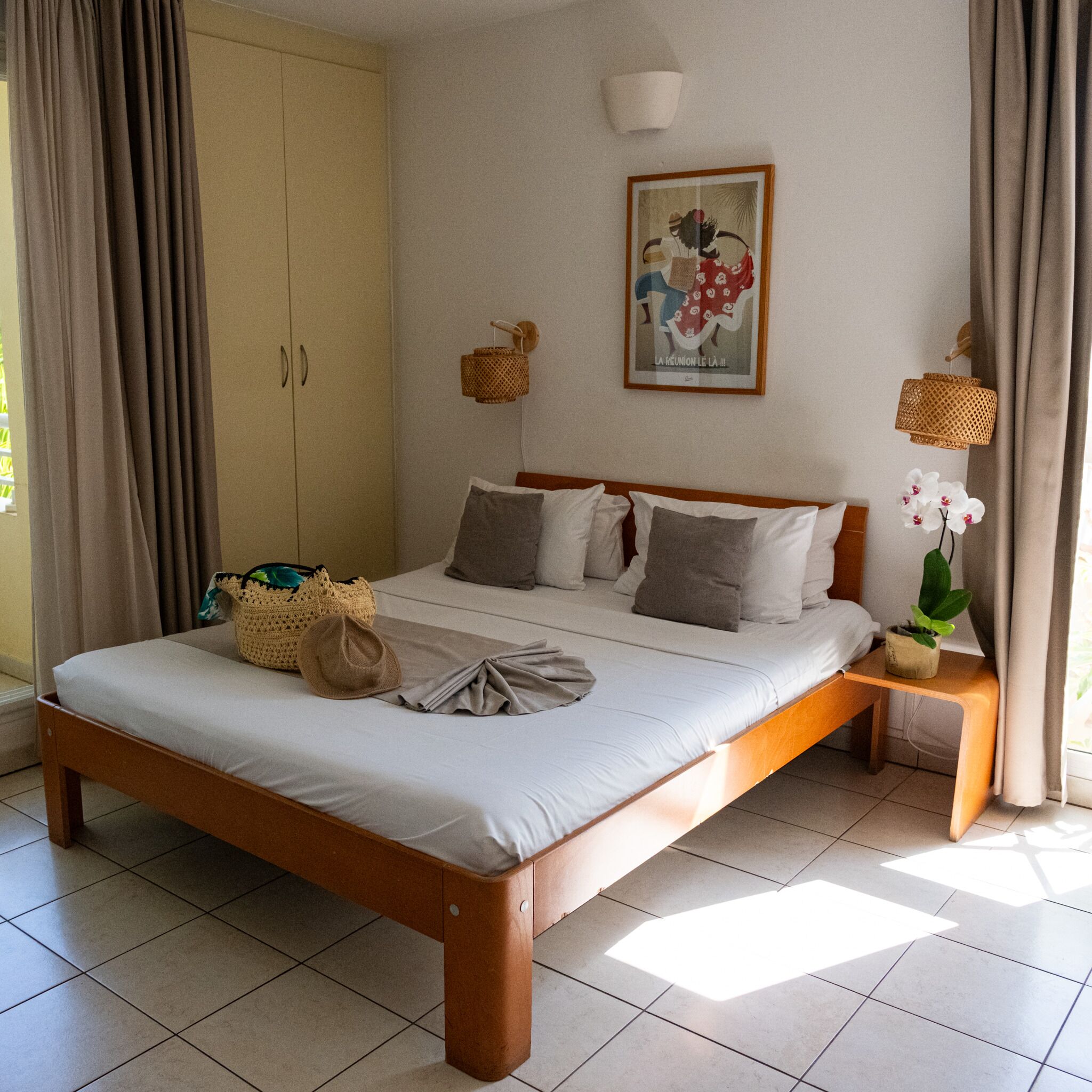 Une chambre à coucher avec un lit, des oreillers, une table de chevet, une lampe fixée au mur, un tableau encadré et une plante en pot.