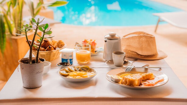 A breakfast table with a variety of food items, a potted plant, and a pool in the background. - Le Kervéguen (Saint-Paul)