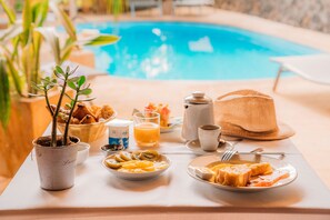Una mesa de desayuno con diversos alimentos, una planta en maceta y una piscina al fondo.