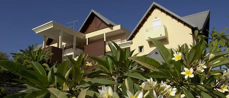A modern two-story house with a balcony and a small garden area with flowering plants.