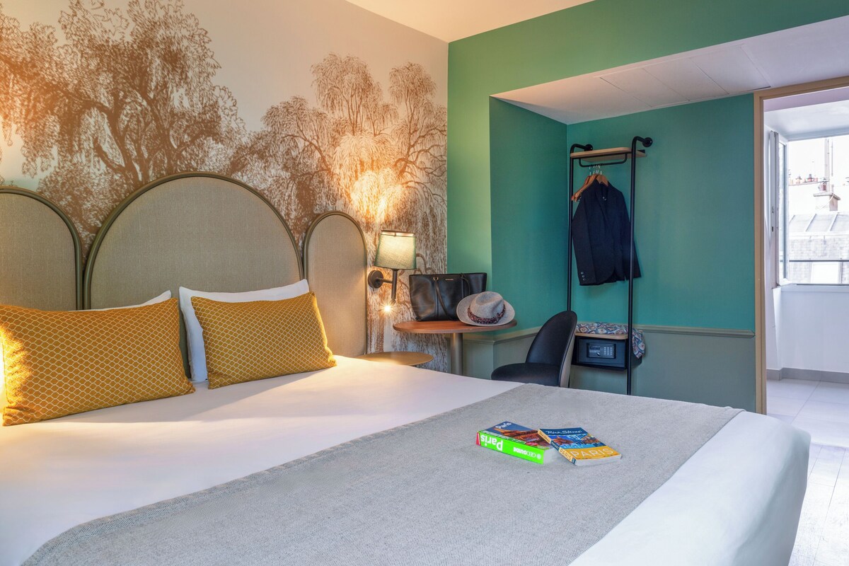 Een hotelkamer met een bed, een nachtkastje, een lamp, een hoed aan de muur en een kapstok met een jas.