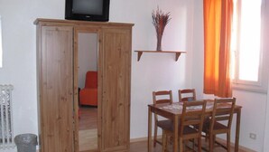 Een kamer met een houten kast, een eettafel met stoelen, een televisie bovenop de kast, een radiator en een raam met oranje gordijnen.