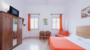 Une chambre avec une armoire en bois, un lit recouvert d’une couverture orange, un bureau avec deux chaises et deux fenêtres ornées de rideaux orange.