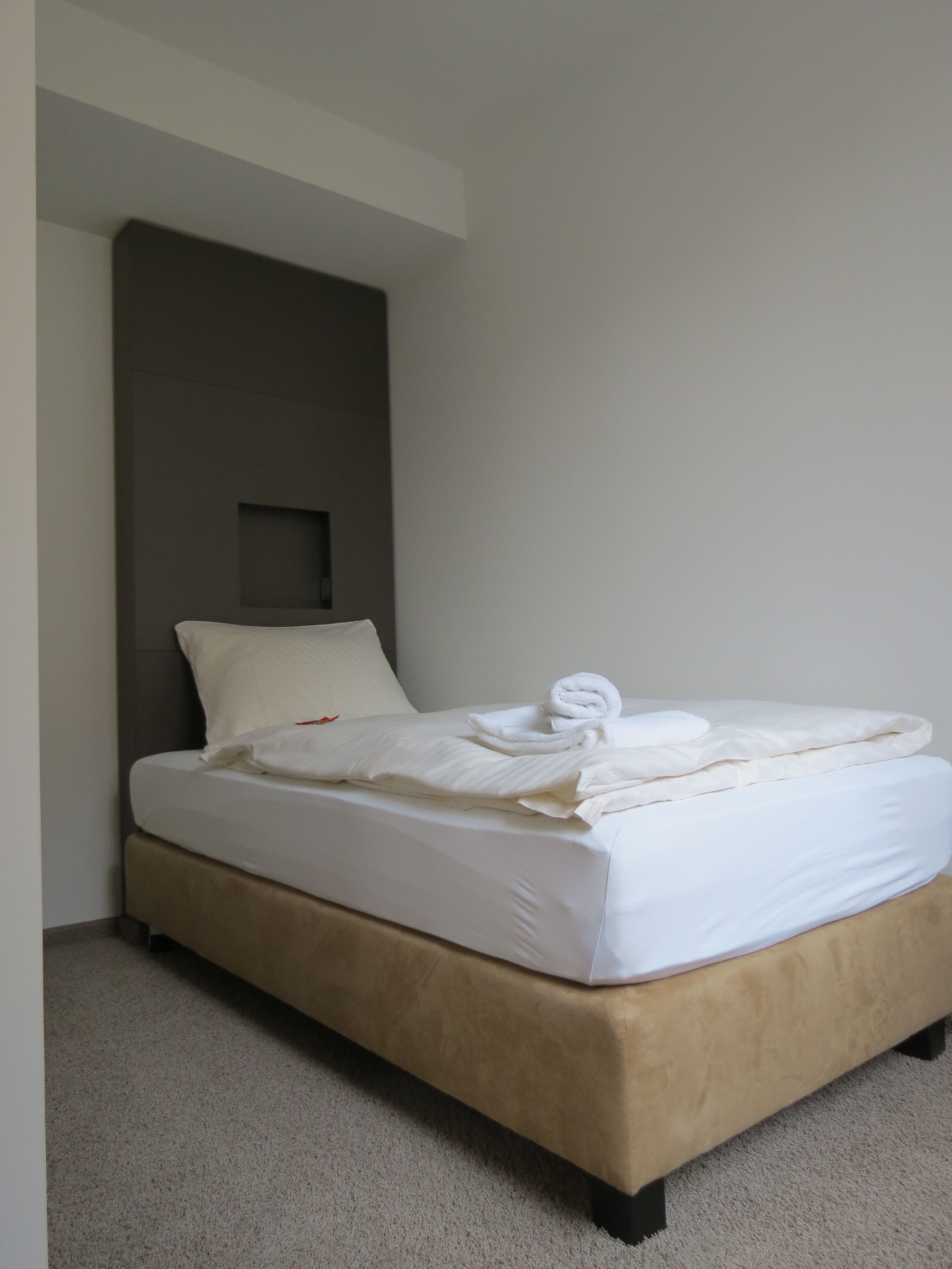 Una cama bien hecha con sábanas blancas y una toalla enrollada, un cabecero tapizado en color beige y suelo alfombrado.