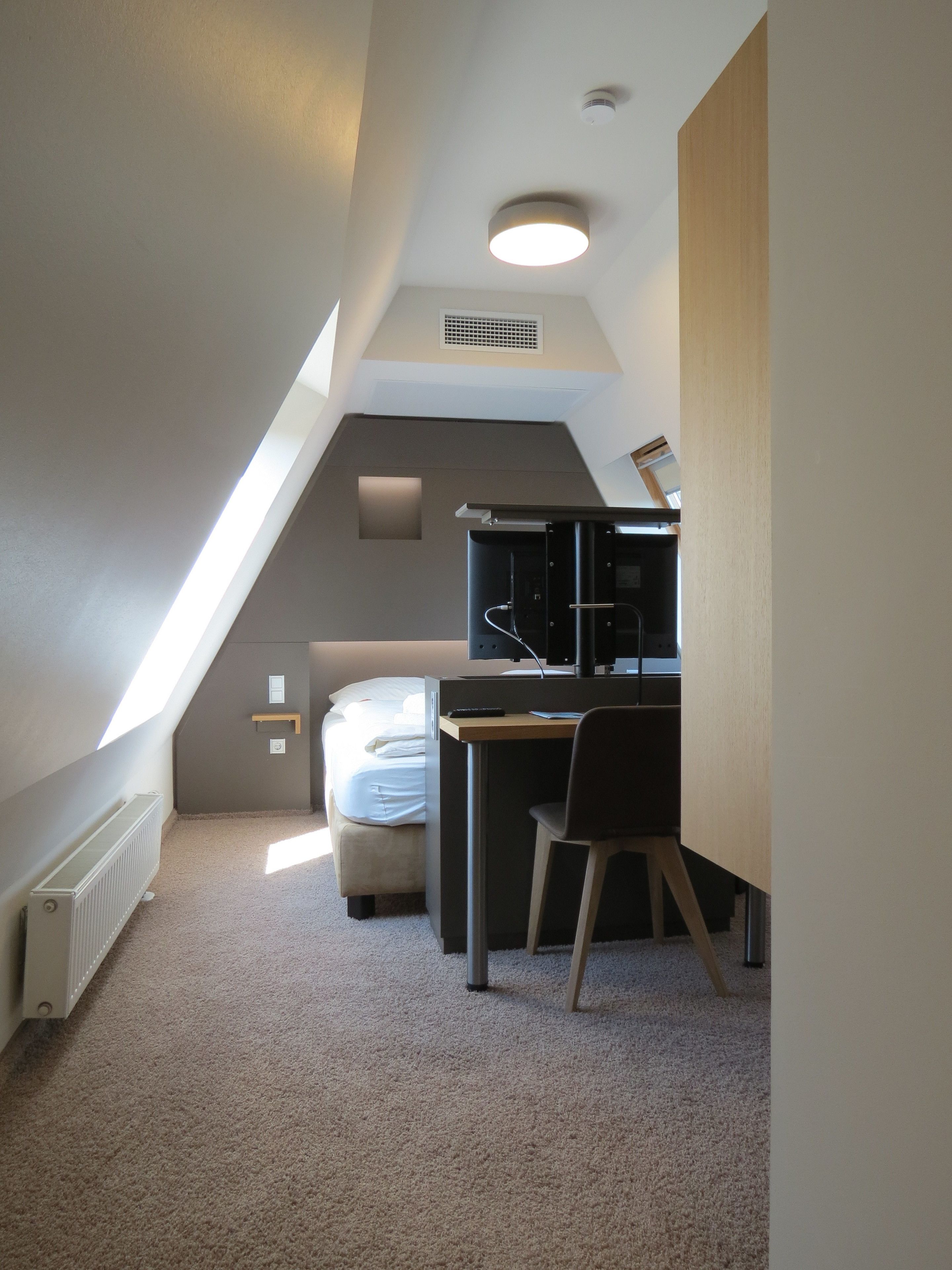 A compact, modern room with a sloped ceiling, a bed, a desk, and a chair.