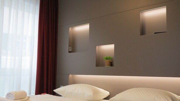 A hotel room with a neatly made bed, a red throw blanket, and two pillows. There are two wall-mounted lights and a small plant on the wall. - Hotel Neuthor (Ulm)