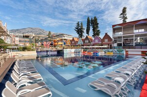 Una piscina con sillones de descanso, un tobogán y edificios coloridos al fondo.