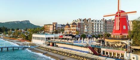 A waterfront hotel with a red windmill, a beach with sun loungers, and a view of the cityscape.