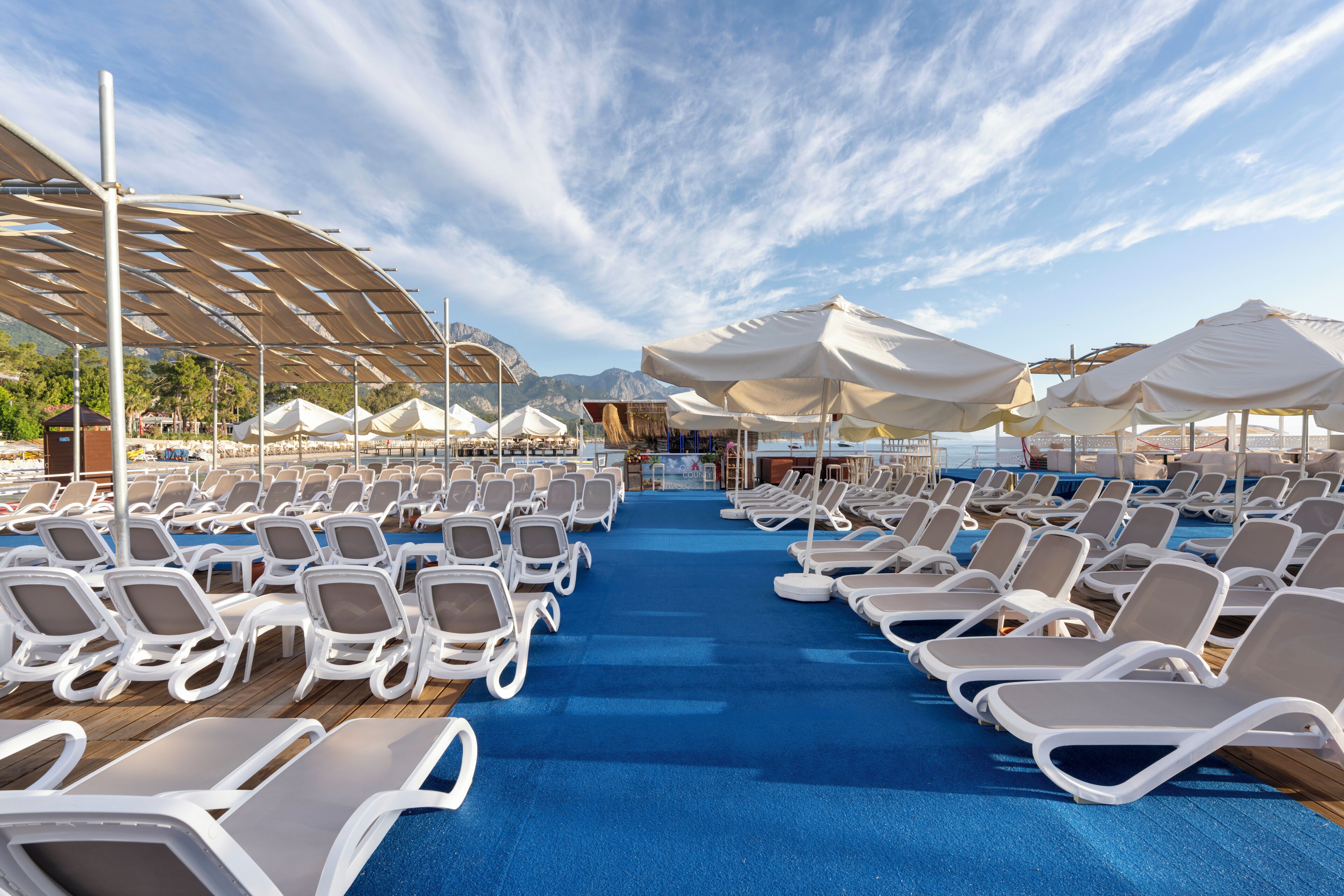 A sun deck with rows of lounge chairs and umbrellas, a wooden deck, and a view of the sea and mountains.