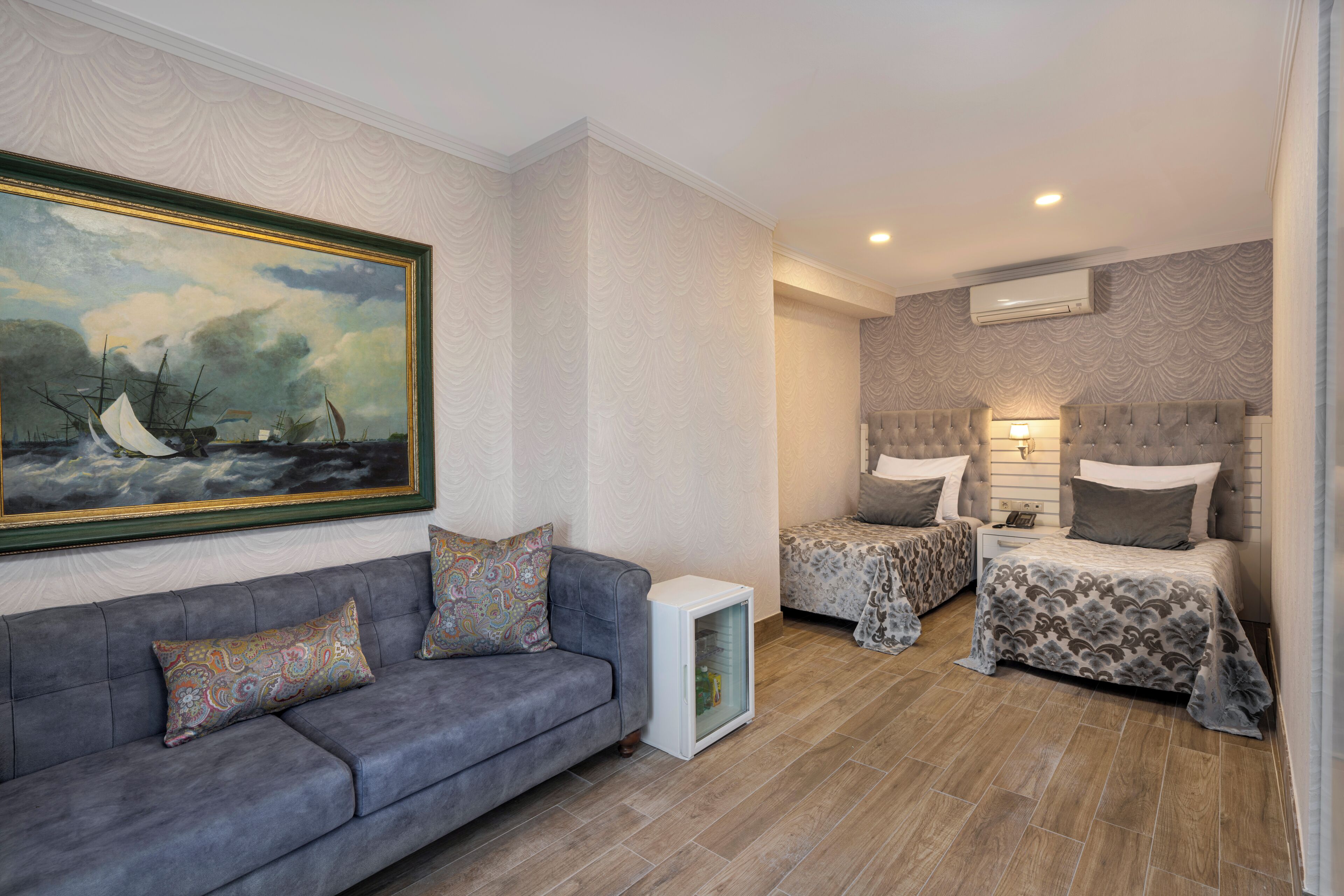 A room with a grey sofa, two beds with patterned bedding, a small cabinet, and a painting on the wall.
