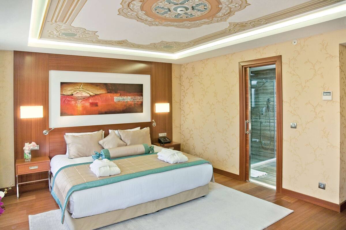A hotel room with a large bed, a painting above the bed, a nightstand with a lamp, and a bathroom door.