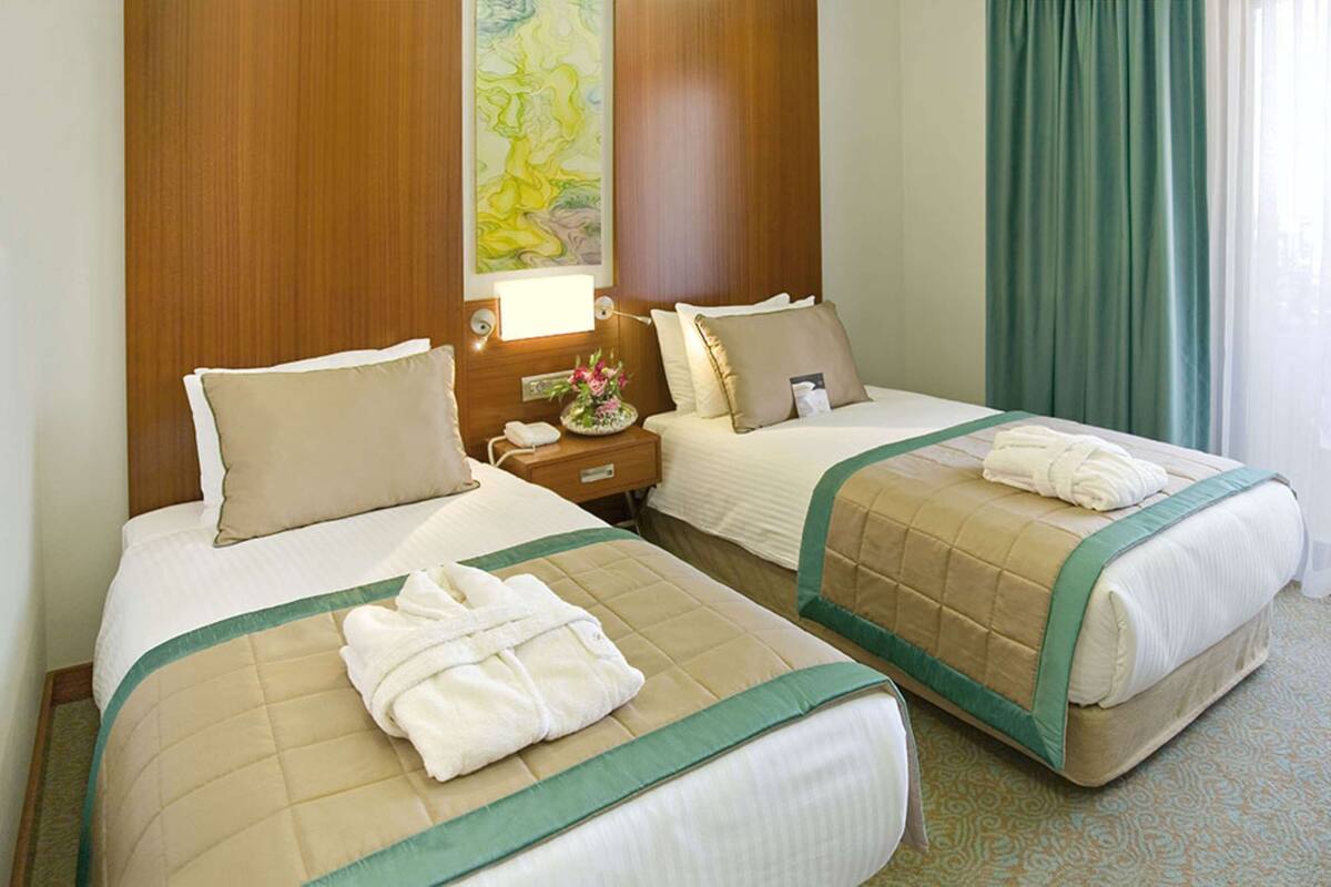 A hotel room with two beds, each with a green and beige bedspread, a nightstand with a lamp, a painting on the wall, and a window with green curtains.