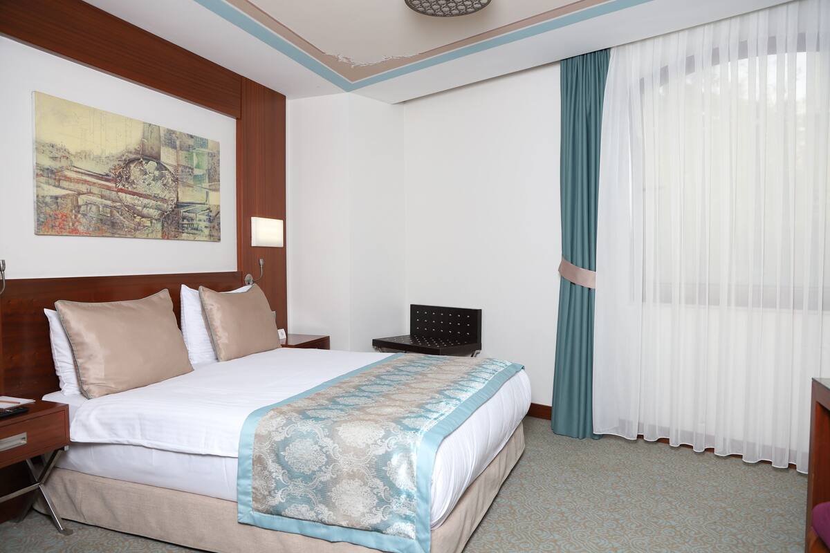 A hotel room with a large bed, a desk, a chair, a painting on the wall, and a window with curtains.