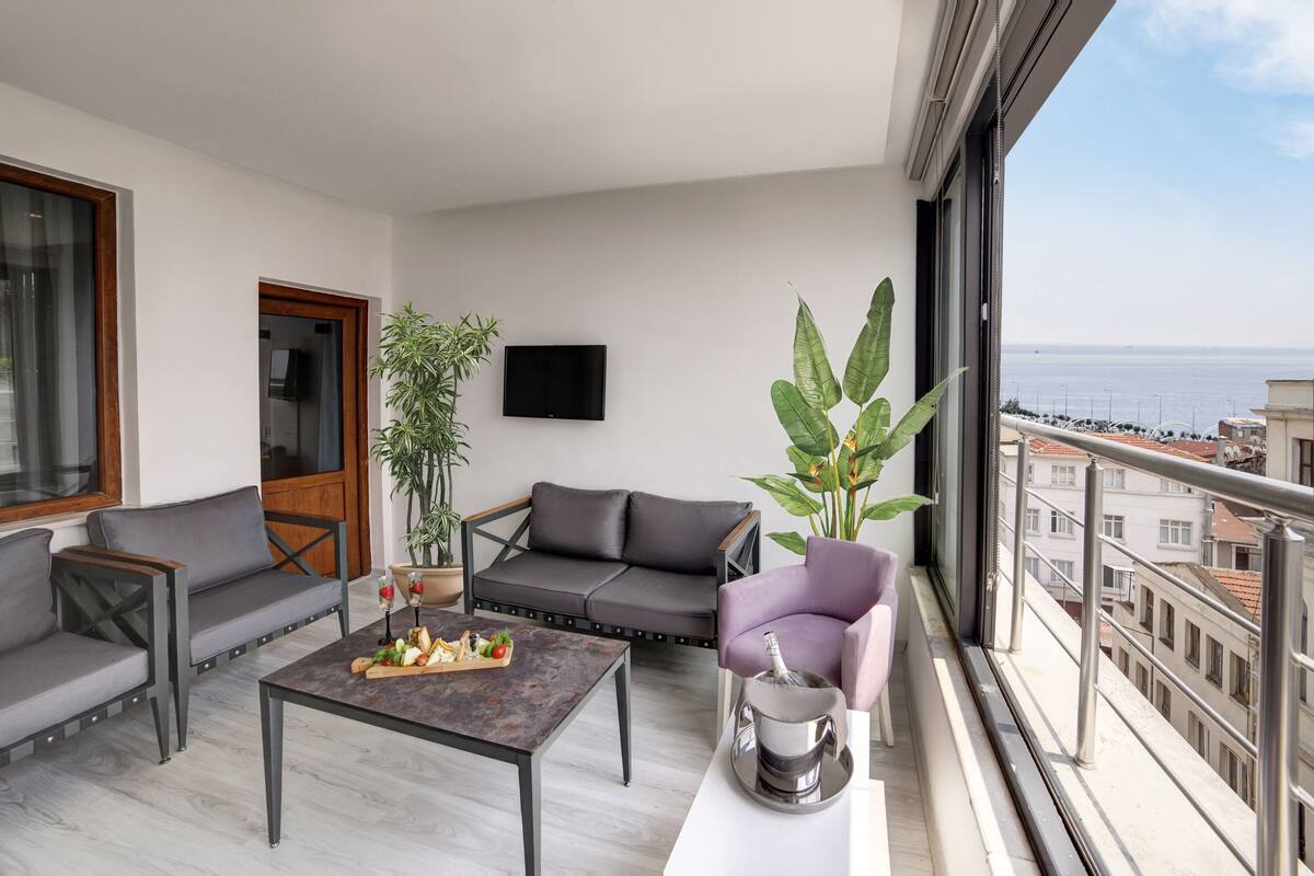 A balcony with a grey sofa, a small table with a bottle and glasses, and a potted plant.