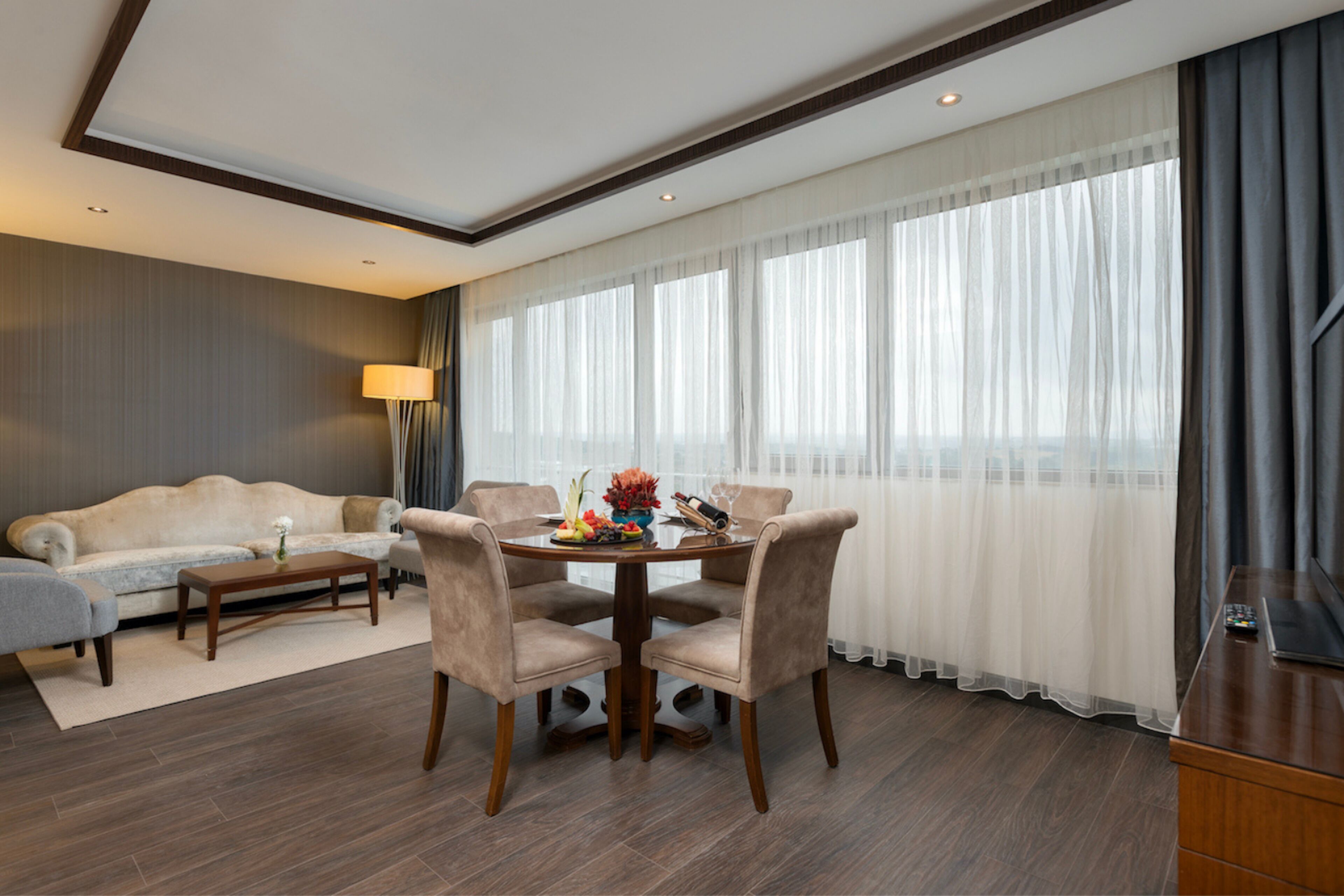 A modern hotel room with a dining table, four chairs, a sofa, a lamp, and a TV.