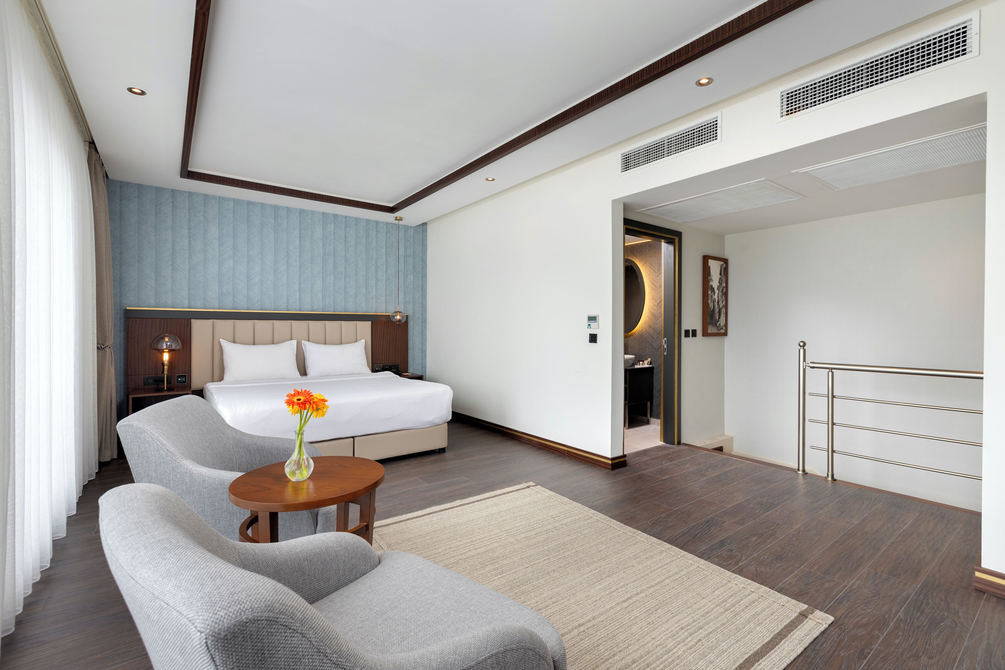 A modern hotel room with a bed, a sofa, a small table with a vase, and a view of the hallway.