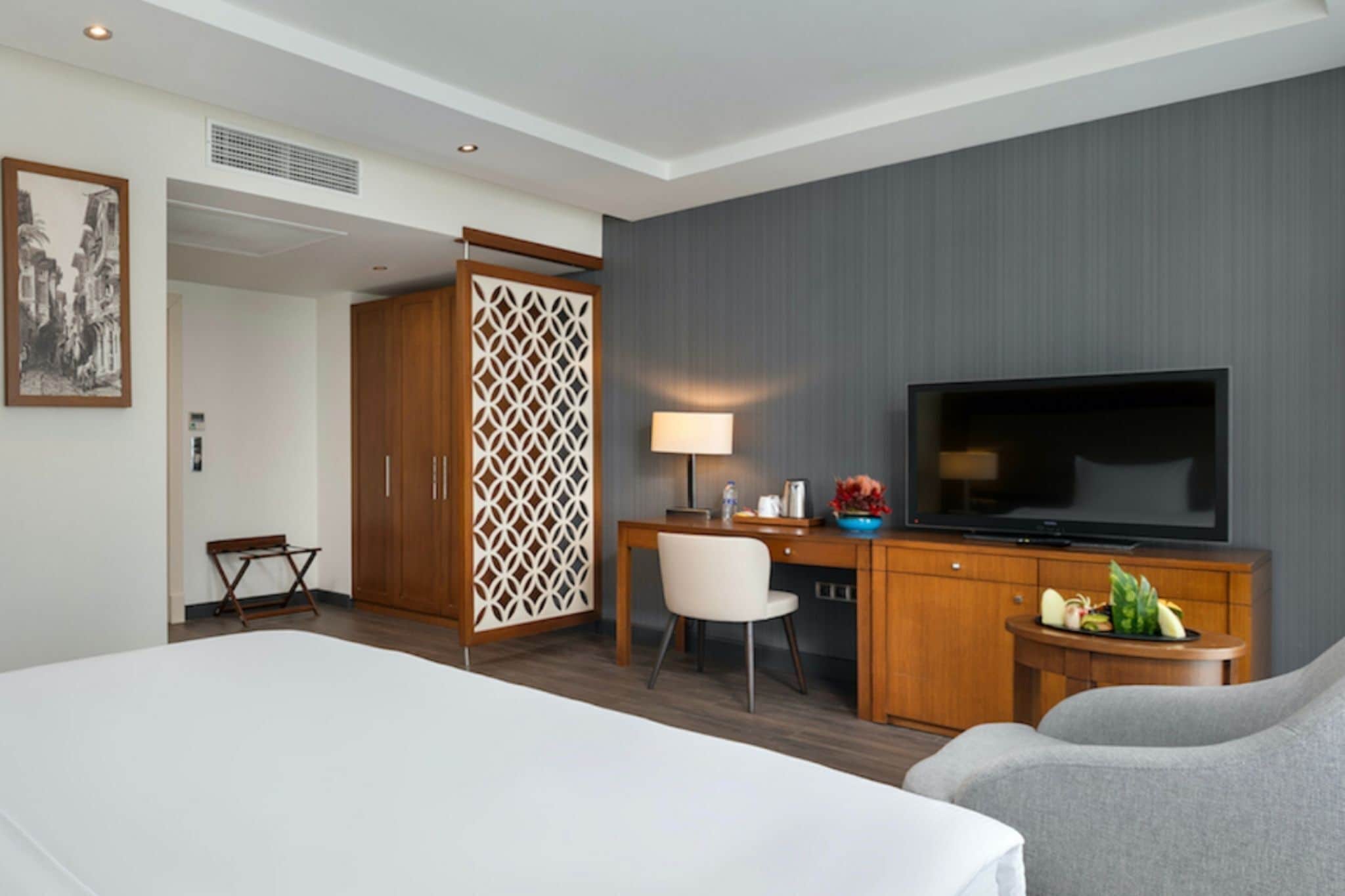 Una habitación de hotel con cama, escritorio con televisor, silla, armario y un separador decorativo.