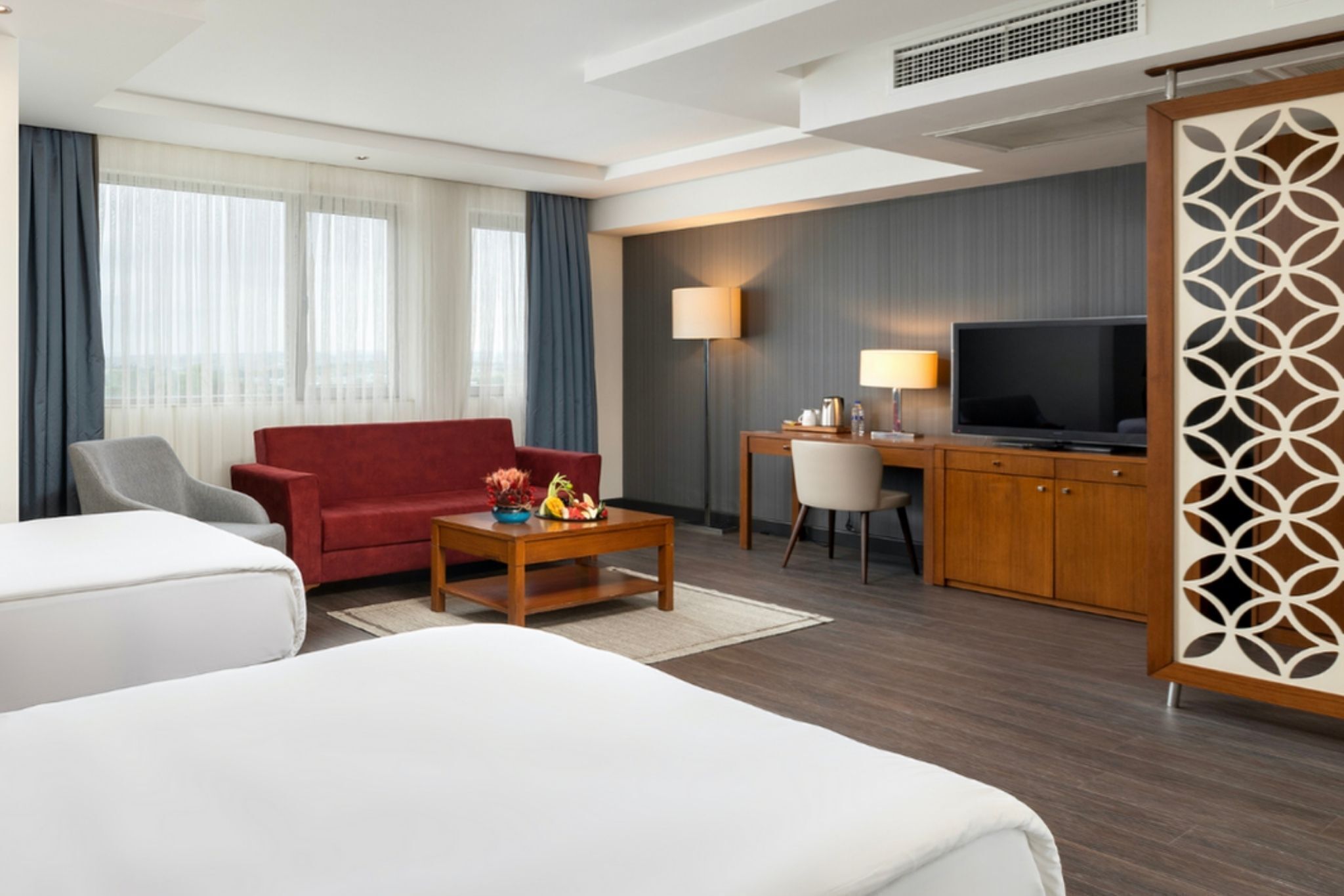 A hotel room with a red sofa, a white sofa, a wooden coffee table with fruit, a desk with a lamp, a TV, and a decorative screen.