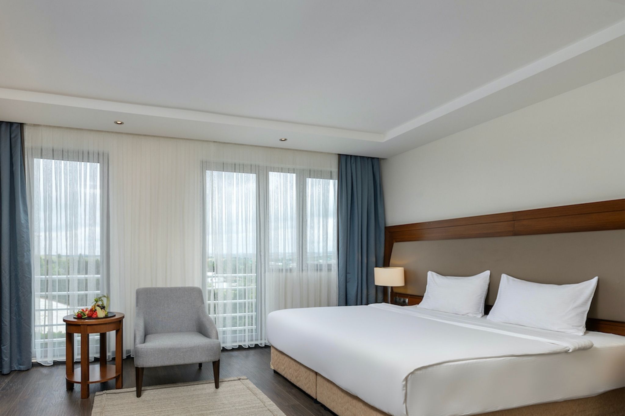 Quarto de hotel com uma cama grande, uma cadeira, uma mesinha com frutas e vista pela janela.
