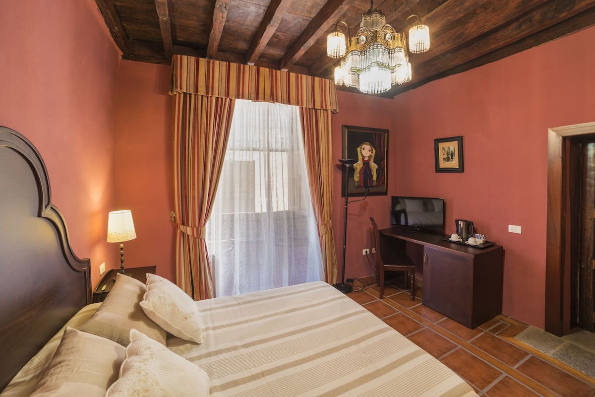 A bedroom with a bed, curtains, a desk, a television, and a chandelier.