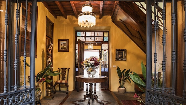 Um corredor imponente com um candelabro, grades de ferro decoradas e uma mesa central com flores.