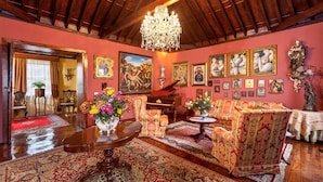 Uma sala de estar tradicional com um lustre imponente, mobiliário ricamente decorado e diversas obras de arte emolduradas nas paredes.