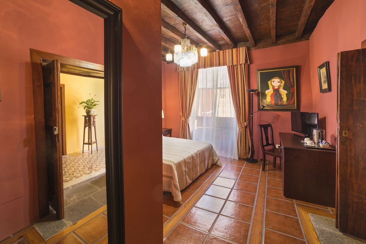 A bedroom with a bed, a desk, a chair, a potted plant, and a painting on the wall.