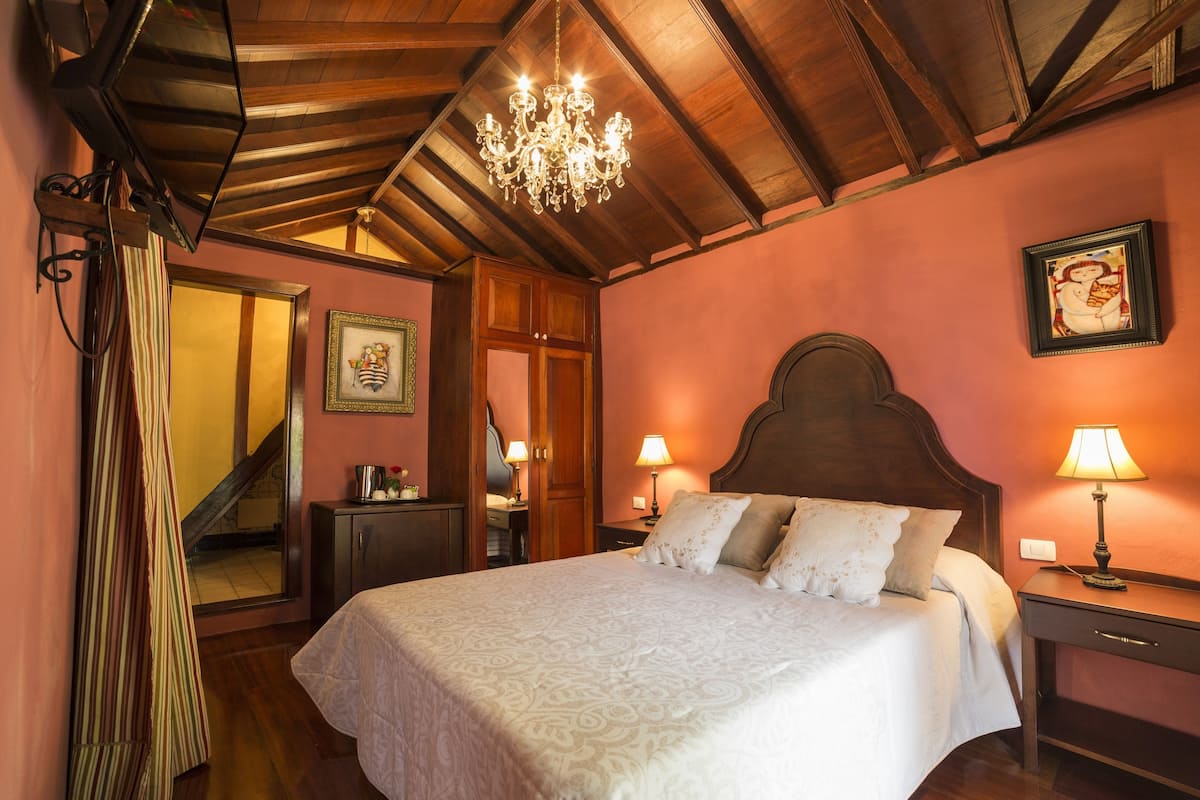 A bedroom with a large bed, wooden ceiling beams, a chandelier, a nightstand with a lamp, and a framed picture on the wall.
