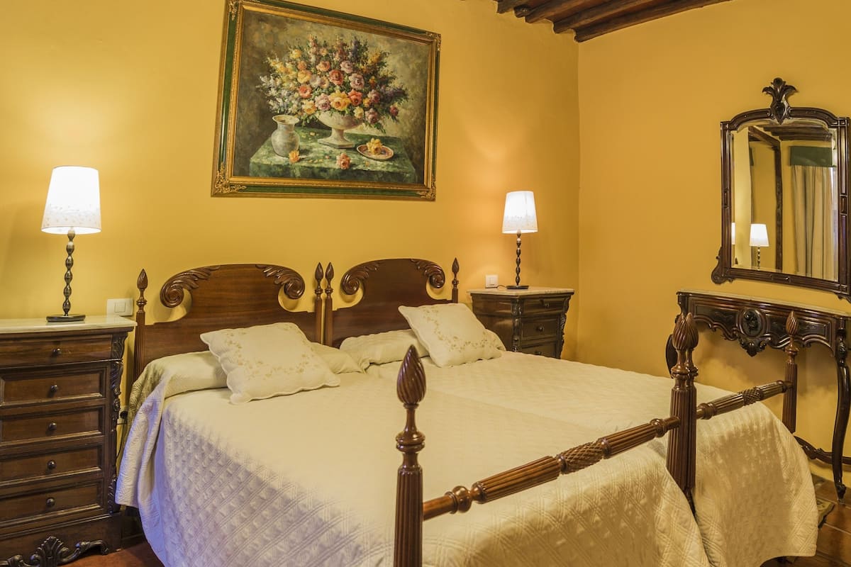 A bedroom with a wooden bed, two bedside tables with lamps, a large mirror, and a floral painting on the wall.