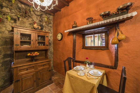 A rustic dining area with a wooden cabinet, a fruit bowl, and a table set for two with plates, glasses, and a vase with a flower. - Hotel Emblematico San Marcos (Icod de los Vinos)