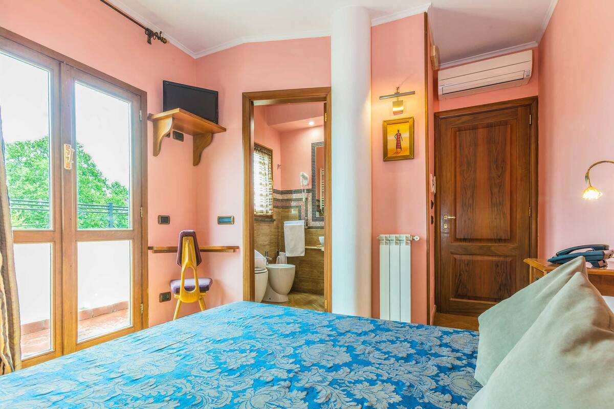 A bedroom with a bed, a chair, a wooden door, and a bathroom visible through an open door.