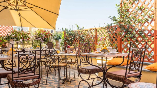 Un café al aire libre con suelo estampado, sillas y mesas de metal bajo un paraguas amarillo grande.