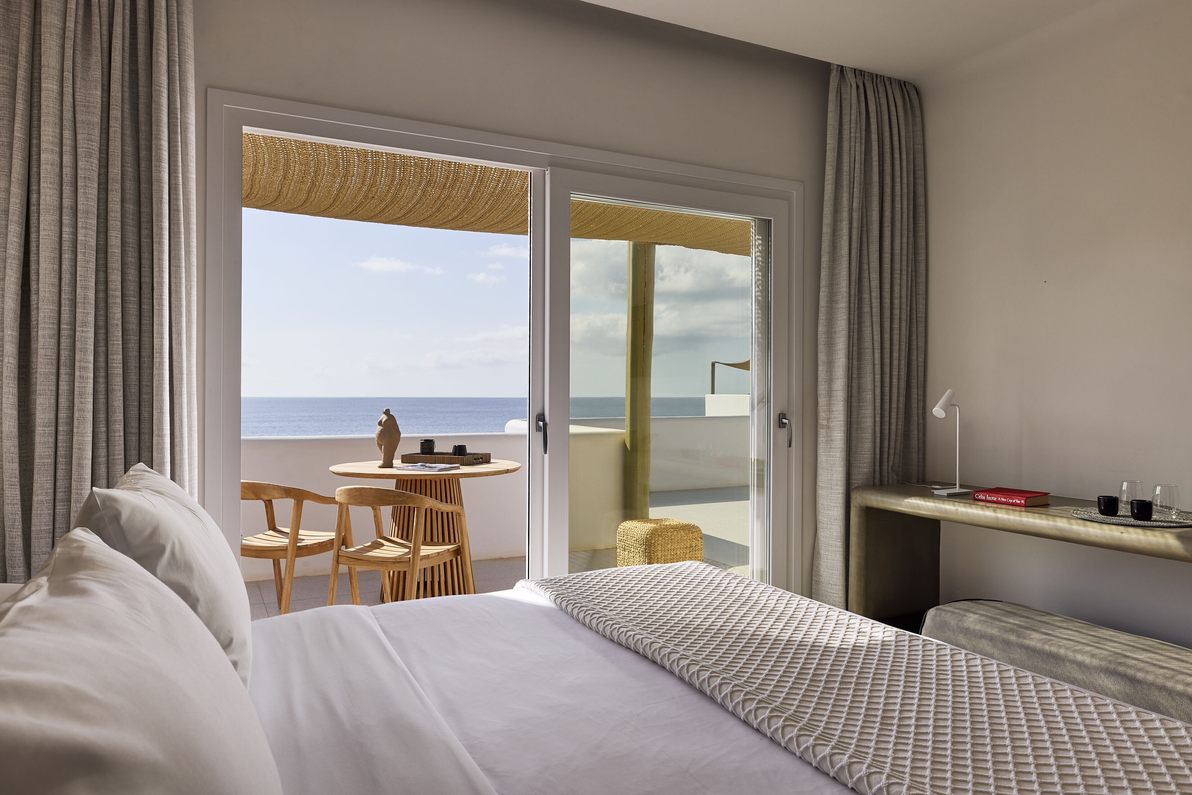 A hotel room with a bed, a small table with chairs, a balcony with a view of the sea, and a shelf with a book and glasses.
