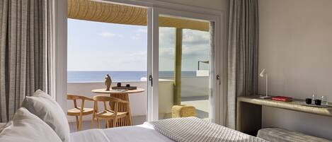 A hotel room with a bed, a small table with chairs, a balcony with a view of the sea, and a shelf with a book and glasses.