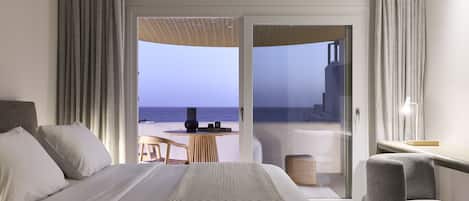 A hotel room with a bed, a chair, a small table, and a view of the sea through a glass door.