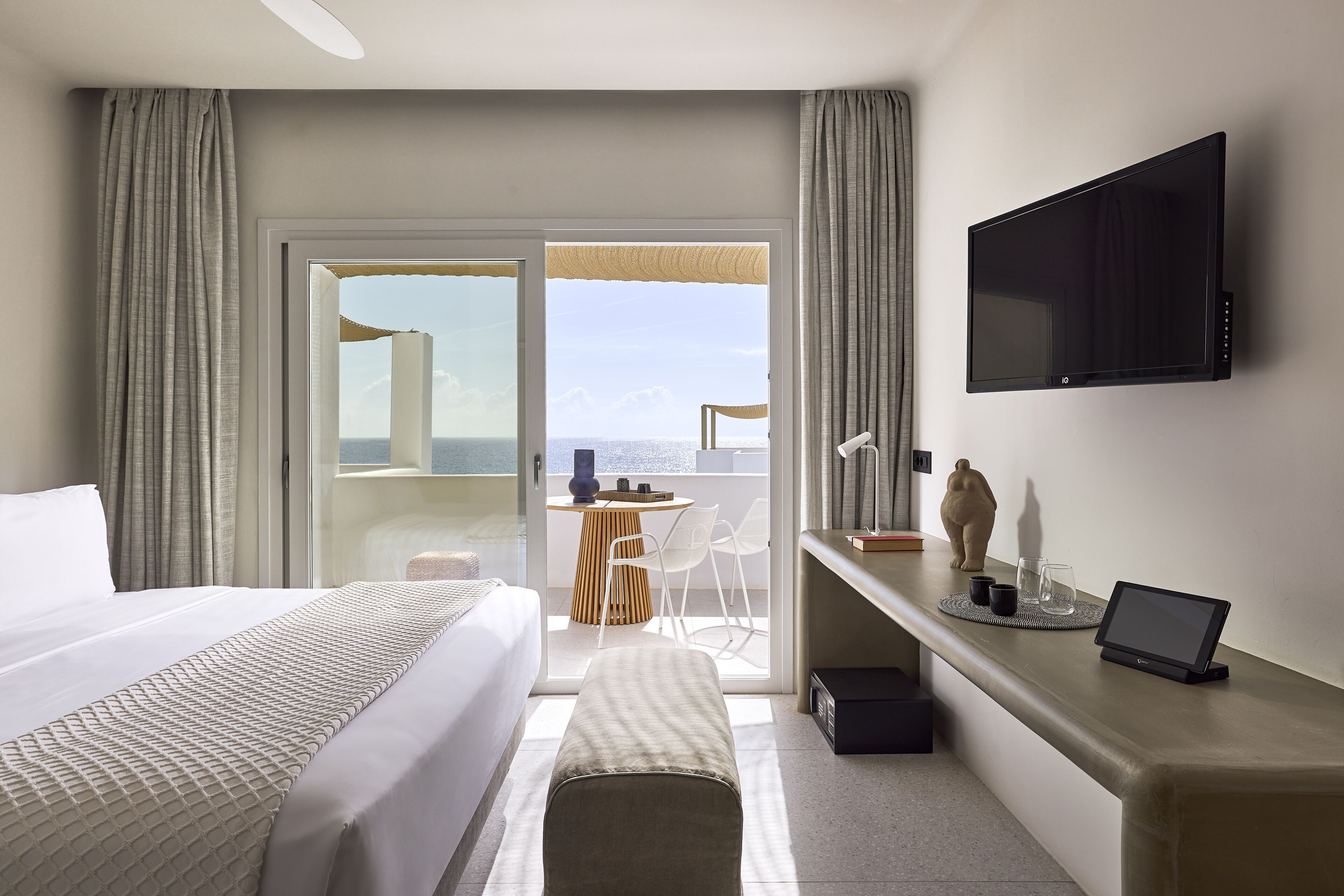 A hotel room with a bed, a desk with a laptop, a TV, and a balcony with a view.