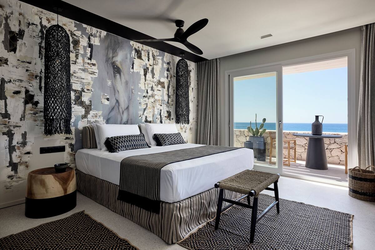 A modern bedroom with a large bed, a ceiling fan, and a view of the sea through large windows.