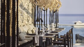 A restaurant with a long table set for dining, a woven hanging light fixture, and a view of the sea. - Artemis Seaside Resort (Milos)